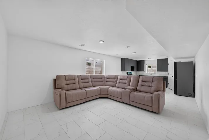 Recessed lighting illuminates a spacious open-concept room featuring tile flooring, a kitchen area with dark cabinetry, and a window providing natural light