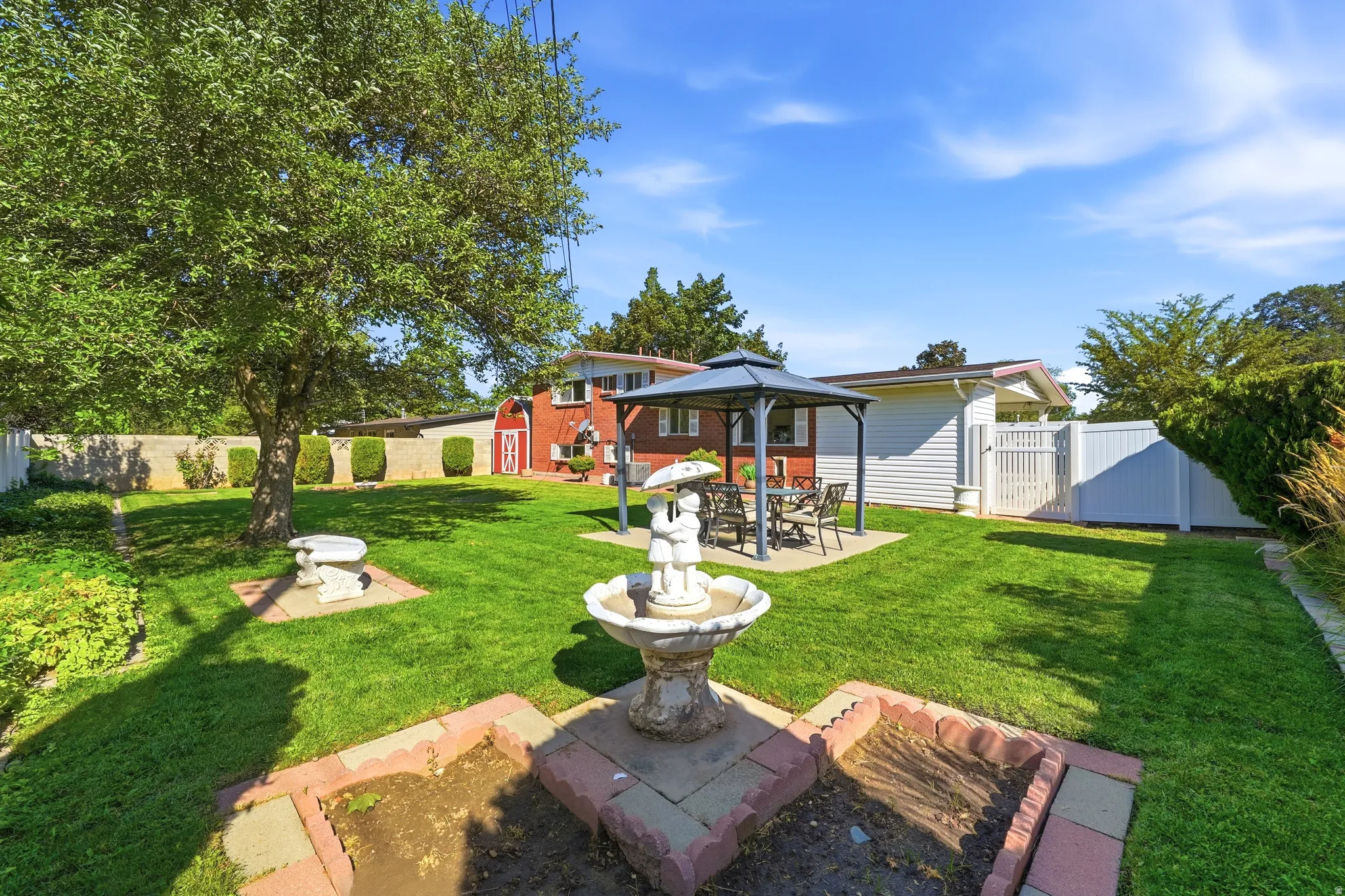 Expansive green lawn, established shade tree, decorative garden fountain, covered patio with outdoor seating, and privacy fencing