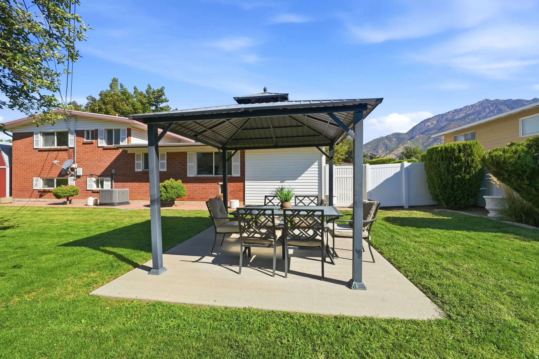 Expansive lawn area with a concrete patio featuring a metal-framed gazebo, surrounded by a white privacy fence, and a mountain backdrop
