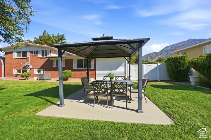 Expansive lawn area with a concrete patio featuring a metal-framed gazebo, surrounded by a white privacy fence, and a mountain backdrop