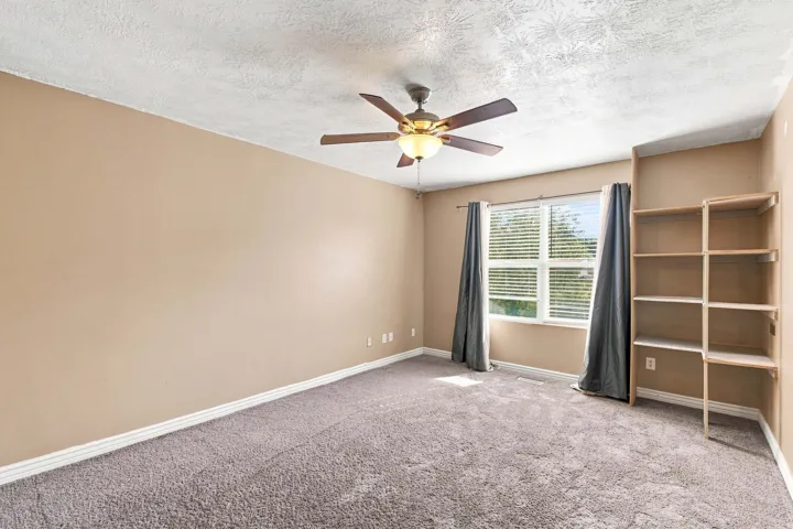 Neutral-toned walls, textured ceiling, ceiling fan with integrated lighting, window with blinds, and built-in shelving