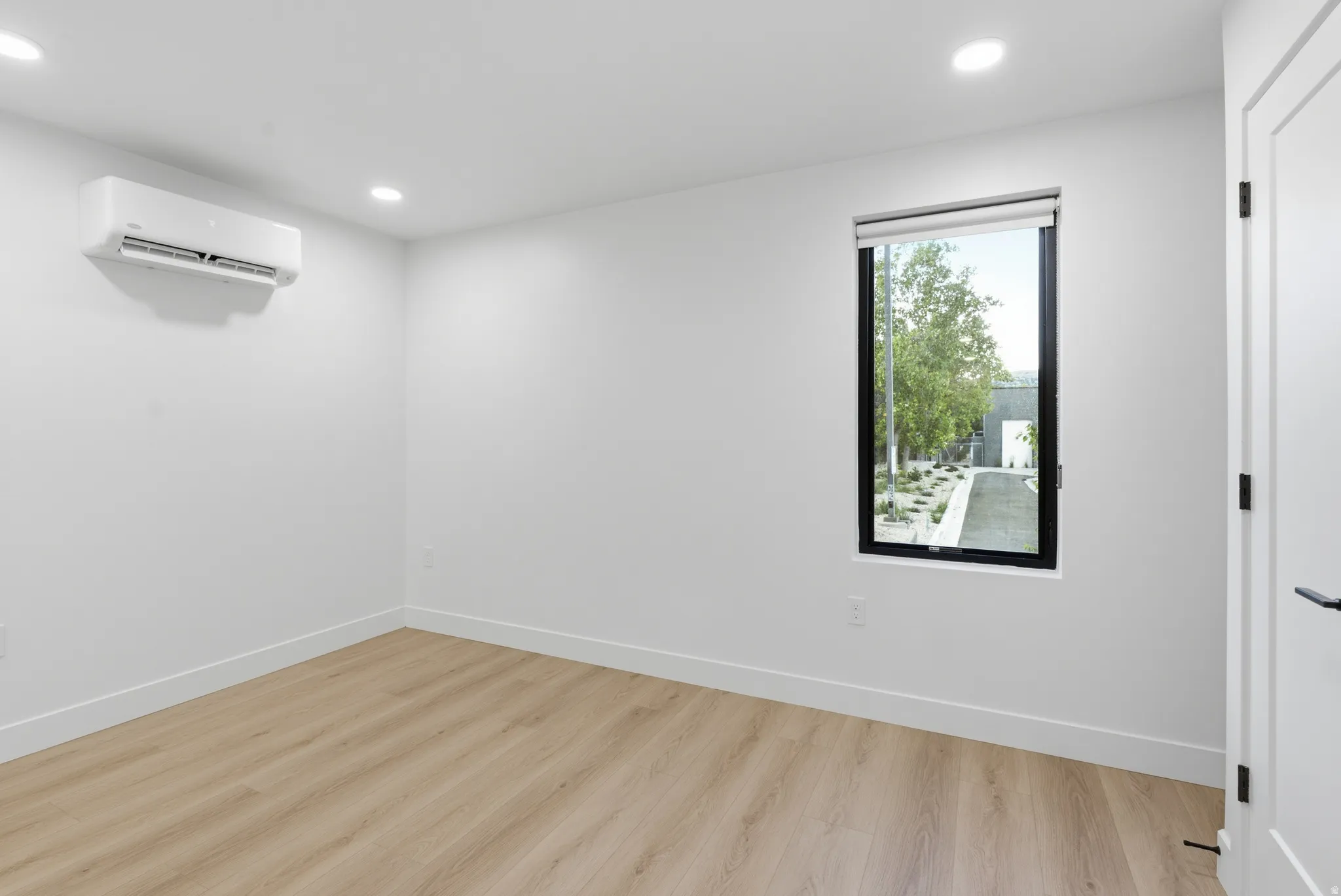Recessed lighting, wood-finish flooring, white walls, black-framed window with roller shade, and wall-mounted air conditioning unit