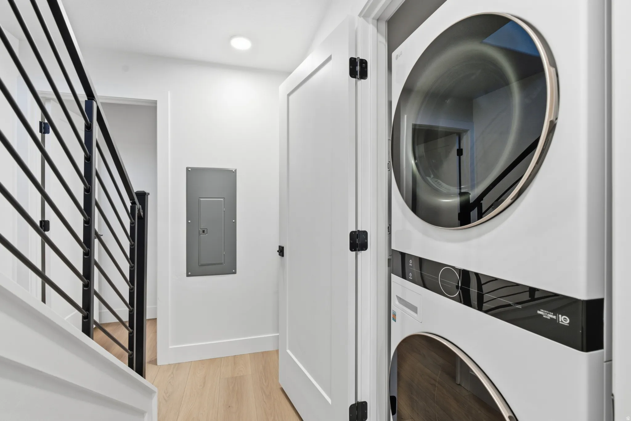 Stacked washer and dryer unit, wood-finish flooring, modern black metal stair railing, recessed ceiling lighting, white interior door with black hardware