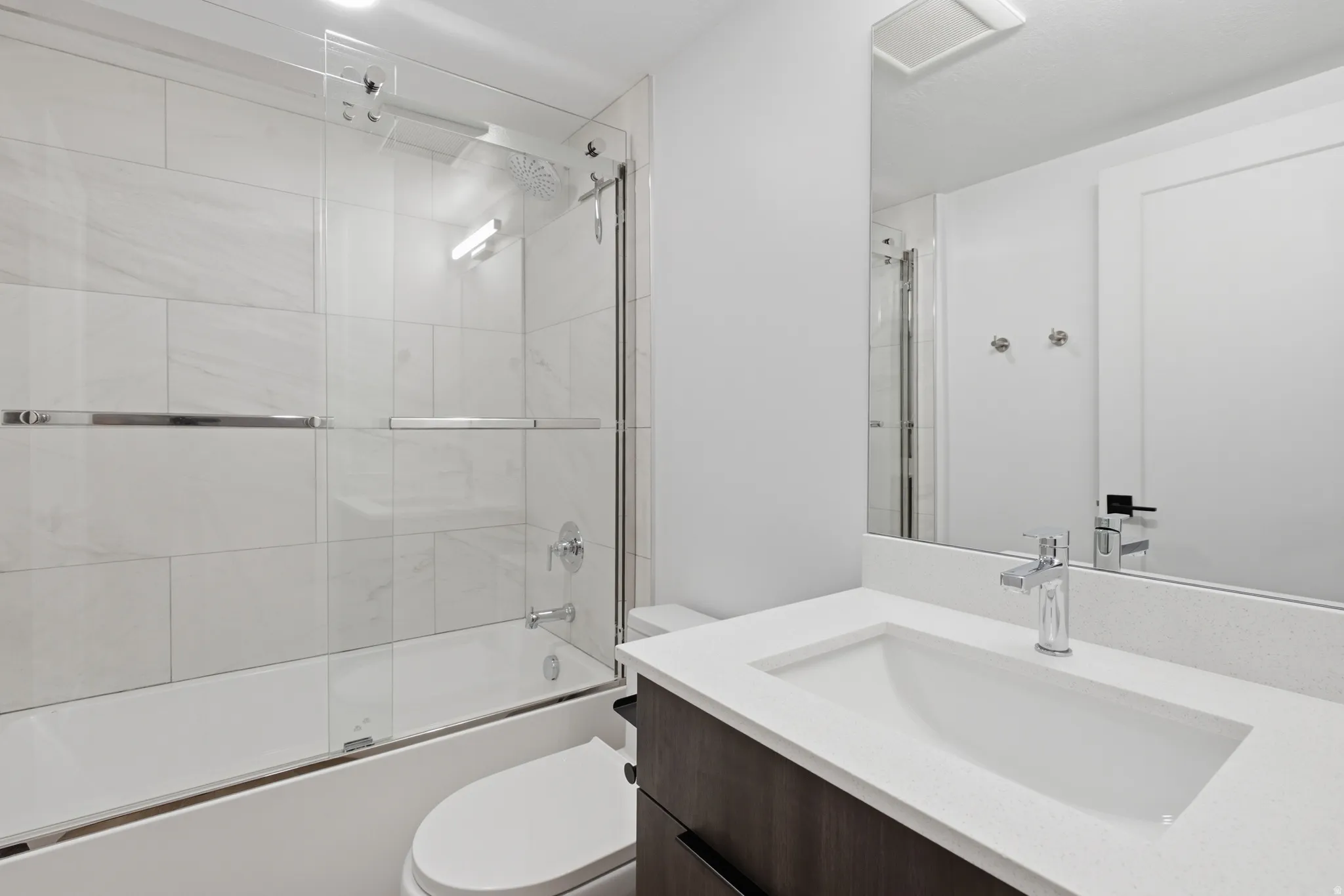 Bathroom featuring a bathtub with a sliding glass enclosure, white rectangular wall tiles, wood-finish vanity with a white countertop, and a rectangular undermount sink