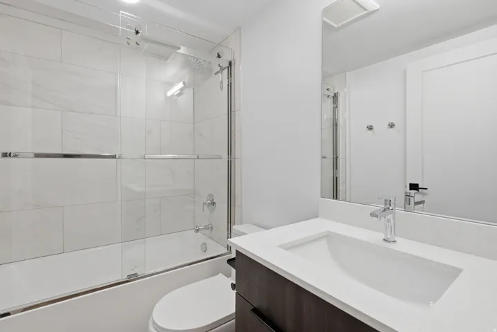 Bathroom featuring a bathtub with a sliding glass enclosure, white rectangular wall tiles, wood-finish vanity with a white countertop, and a rectangular undermount sink