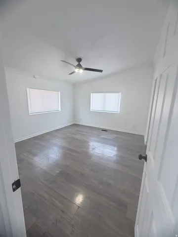 Spacious room featuring wood-finish flooring, vaulted ceiling, dual windows with blinds, crown molding, and a ceiling fan with light
