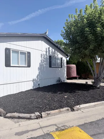 White vertical siding exterior with dark shutters, dark roof, landscaped bed with dark mulch, and mature perimeter trees