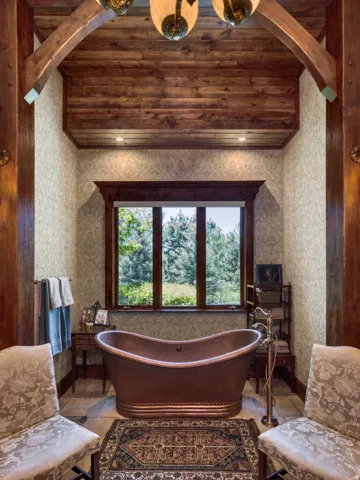 Freestanding copper bathtub with a floor-mounted faucet, wood-paneled ceiling, exposed wood beams, and a wood-trimmed window