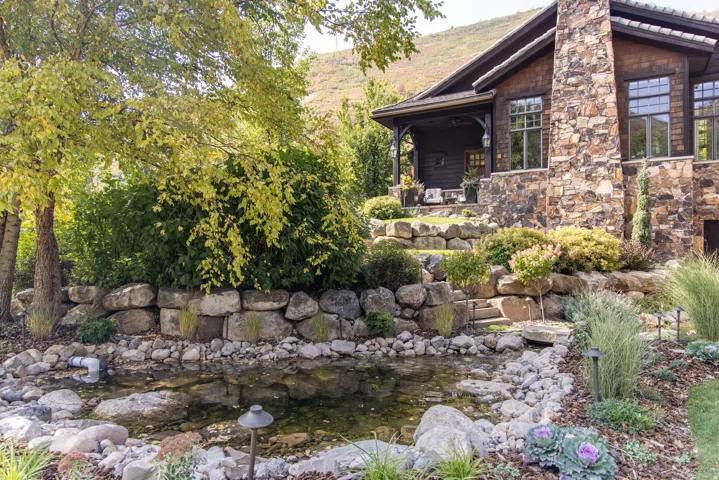 Stone-clad exterior with multiple windows, tiered landscaping featuring rock walls, a tranquil pond, and mature trees