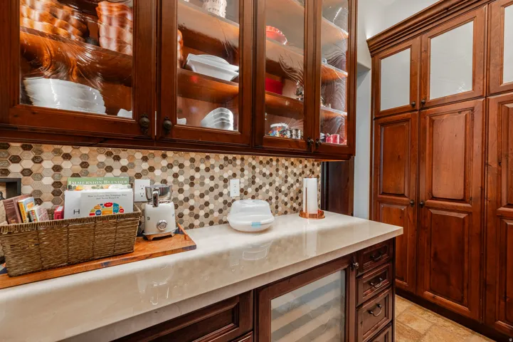 Custom wood cabinetry with glass fronts, integrated under-cabinet lighting, hexagonal mosaic tile backsplash, solid surface countertop, and built-in beverage refrigerator