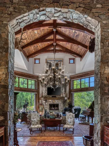 Stone archway framing a grand living space with a vaulted wood-beam ceiling, a substantial stone fireplace, expansive windows, and a large chandelier