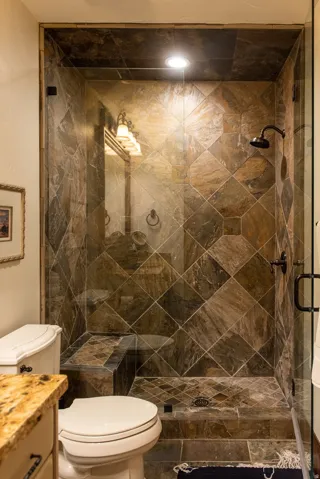 Walk-in shower with natural stone tile, glass enclosure, built-in bench, and dark bronze fixtures