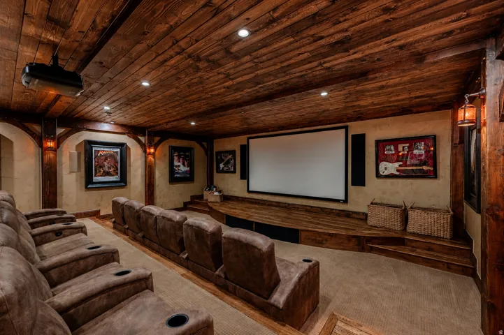 Wood-paneled ceiling with recessed lighting, large projection screen, multi-tiered seating with integrated cup holders, wood-finish stage, and wall-mounted sconces