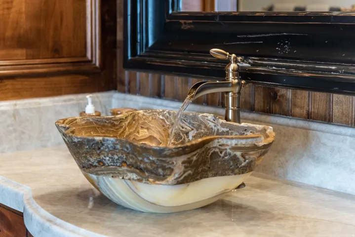 Vessel sink with natural stone finish, waterfall faucet, light-toned countertop, wood paneling, and dark-framed mirror