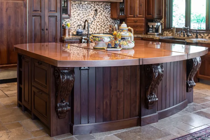 Curved kitchen island with a copper countertop, dark wood cabinetry, and carved corbel accents
