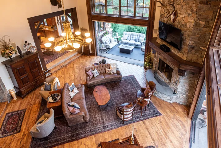 Expansive living space featuring wood-finish flooring, a stone fireplace with a substantial timber mantel, and a large chandelier