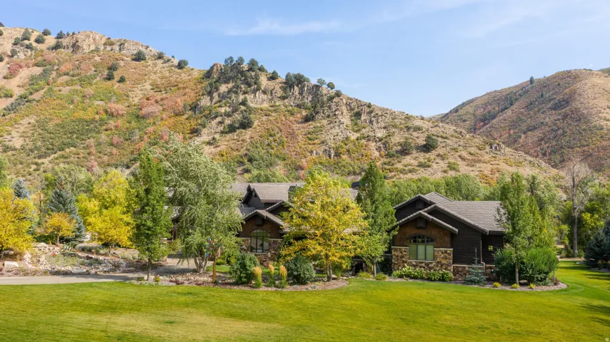 Expansive green lawn with mature trees and a stone-clad home featuring dark siding and a prominent roofline, set against a mountainous backdrop