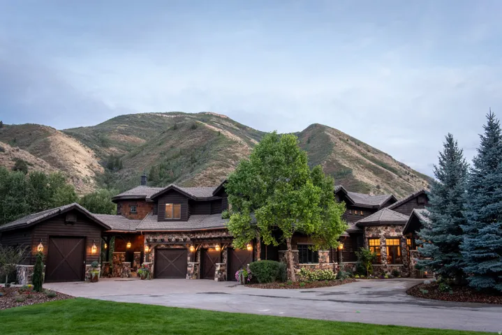 Stone and wood-sided exterior with multiple garage bays, integrated outdoor lighting, mature trees, and mountain scenery