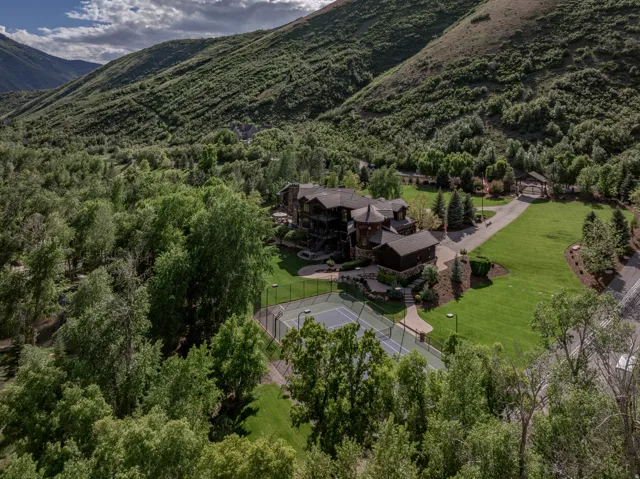 Expansive property featuring a multi-story residence with a dark roof and stone accents, a full-size tennis court, a long paved driveway, substantial green lawns, and mature trees