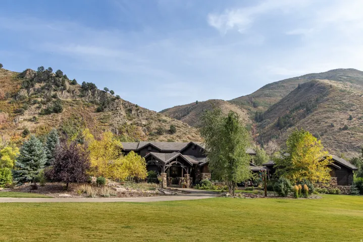 Expansive green lawn with mature trees, stone and timber-clad exterior, covered entry, and a mountain backdrop