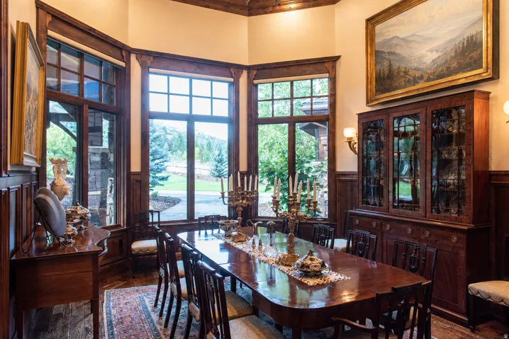 Elegant formal dining room showcasing rich wood cabinetry with glass-front display cases, a polished wood-finish dining table, and multiple multi-pane windows framing natural landscape views