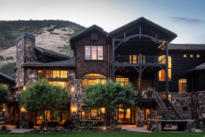 Stunning residence featuring a multi-level stone and timber exterior with peaked gable rooflines, expansive windows with warm interior lighting, and a covered front porch with decorative timber framing
