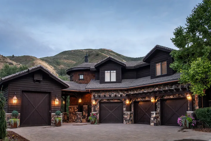 Stone and wood siding exterior with multiple dormers, multi-car garage with custom wood doors, and decorative outdoor lighting