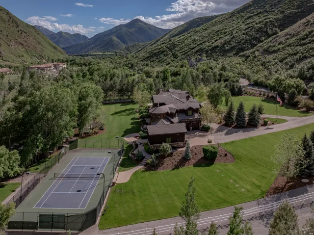 Expansive property featuring a private tennis court with surrounding fencing, a substantial residence with multiple gables and dormers, a long paved driveway, and meticulously maintained lawns