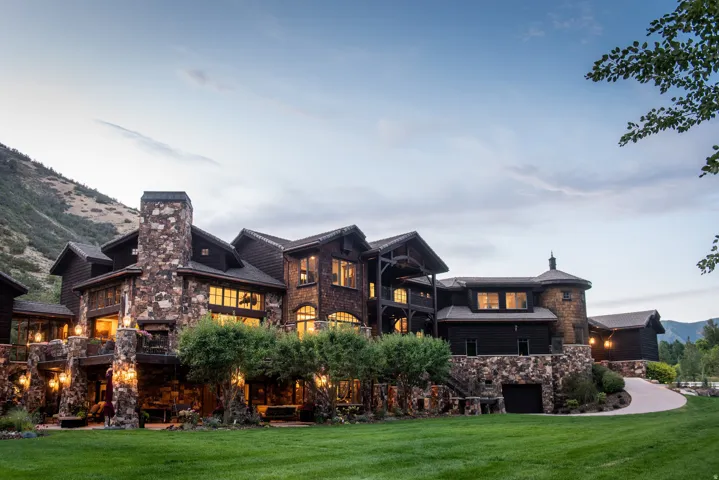 Multi-level stone and wood exterior with multiple windows, prominent stone chimney, and a curved driveway leading to a garage entrance