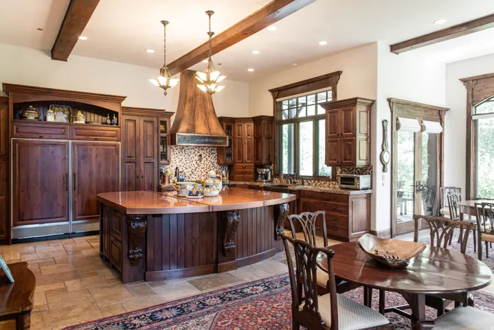 Expansive kitchen featuring wood-finish cabinetry, an oversized island with a copper-finish countertop, a custom range hood, patterned tile backsplash, and exposed ceiling beams