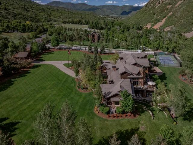 Expansive estate featuring a multi-level stone and wood-sided residence, a private tennis court, a long paved driveway, and mature landscaping with manicured lawns