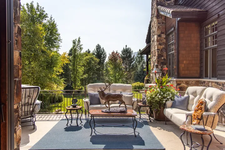 Expansive outdoor patio with a stone facade, shingle siding, built-in wall lantern, and a wrought iron railing overlooking mature trees