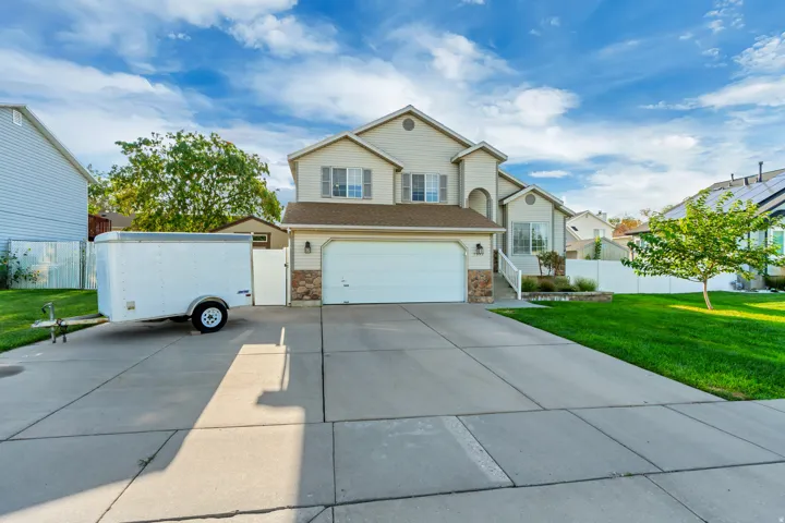 Two-car garage with white overhead doors, expansive concrete driveway with clean geometric sections, lush manicured lawn, and two-story colonial-style home featuring stone and siding exterior with multiple windows and pitched roofline