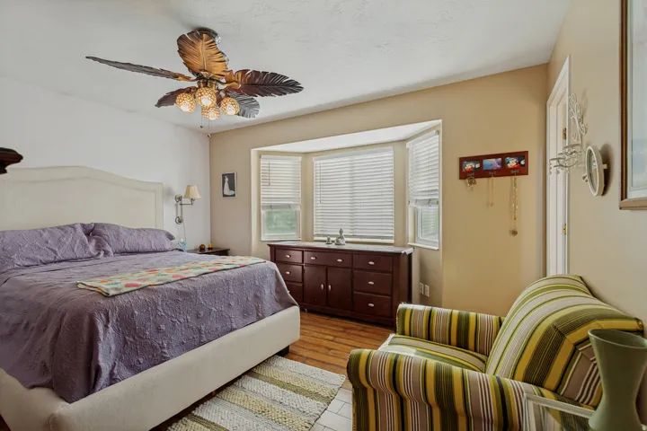 Bright secondary bedroom showcasing hardwood flooring, a ceiling fan with decorative blades, and large multi-pane windows with plantation shutters