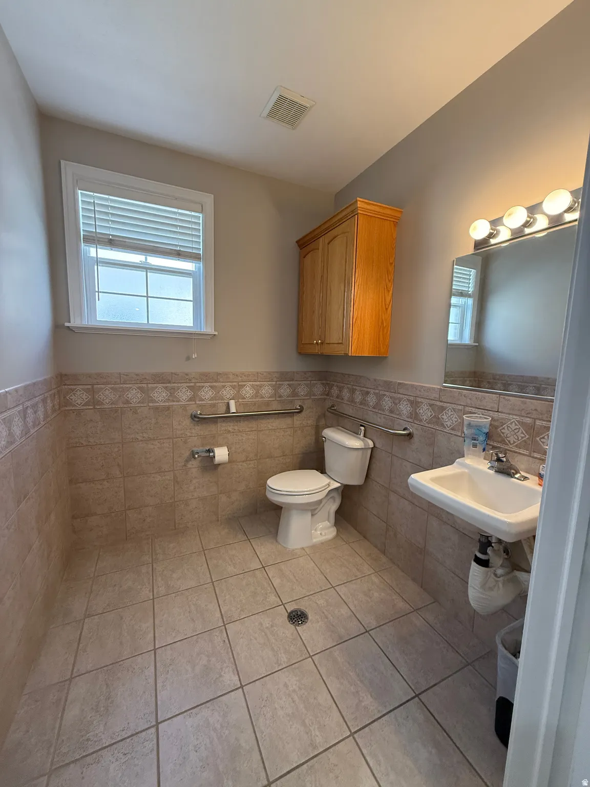Tiled flooring with corresponding wainscoting, wall-mounted vanity sink, white window with blinds, and built-in wood-finish cabinetry