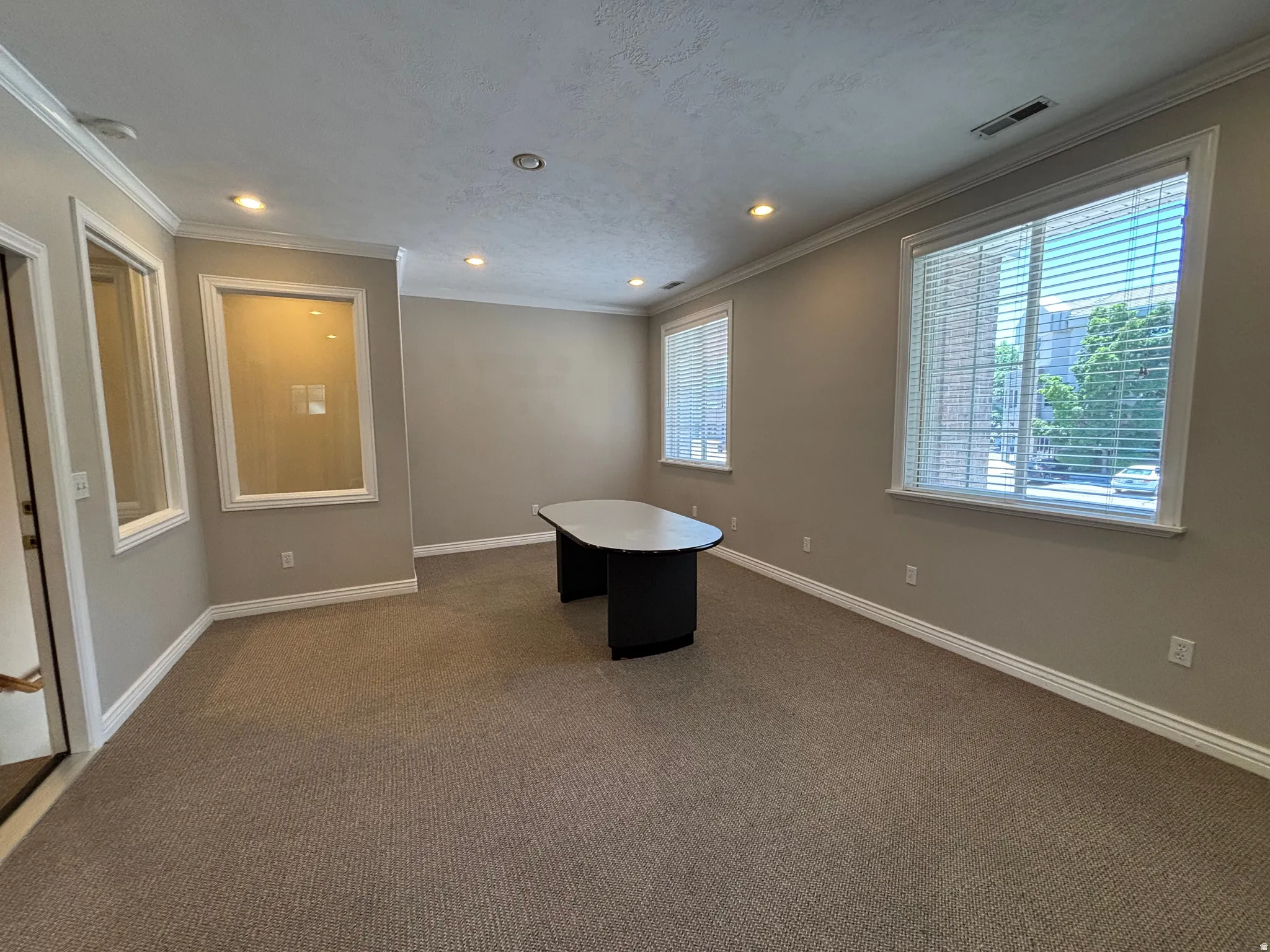 Recessed lighting illuminates a carpeted room featuring multiple windows with blinds and crown molding