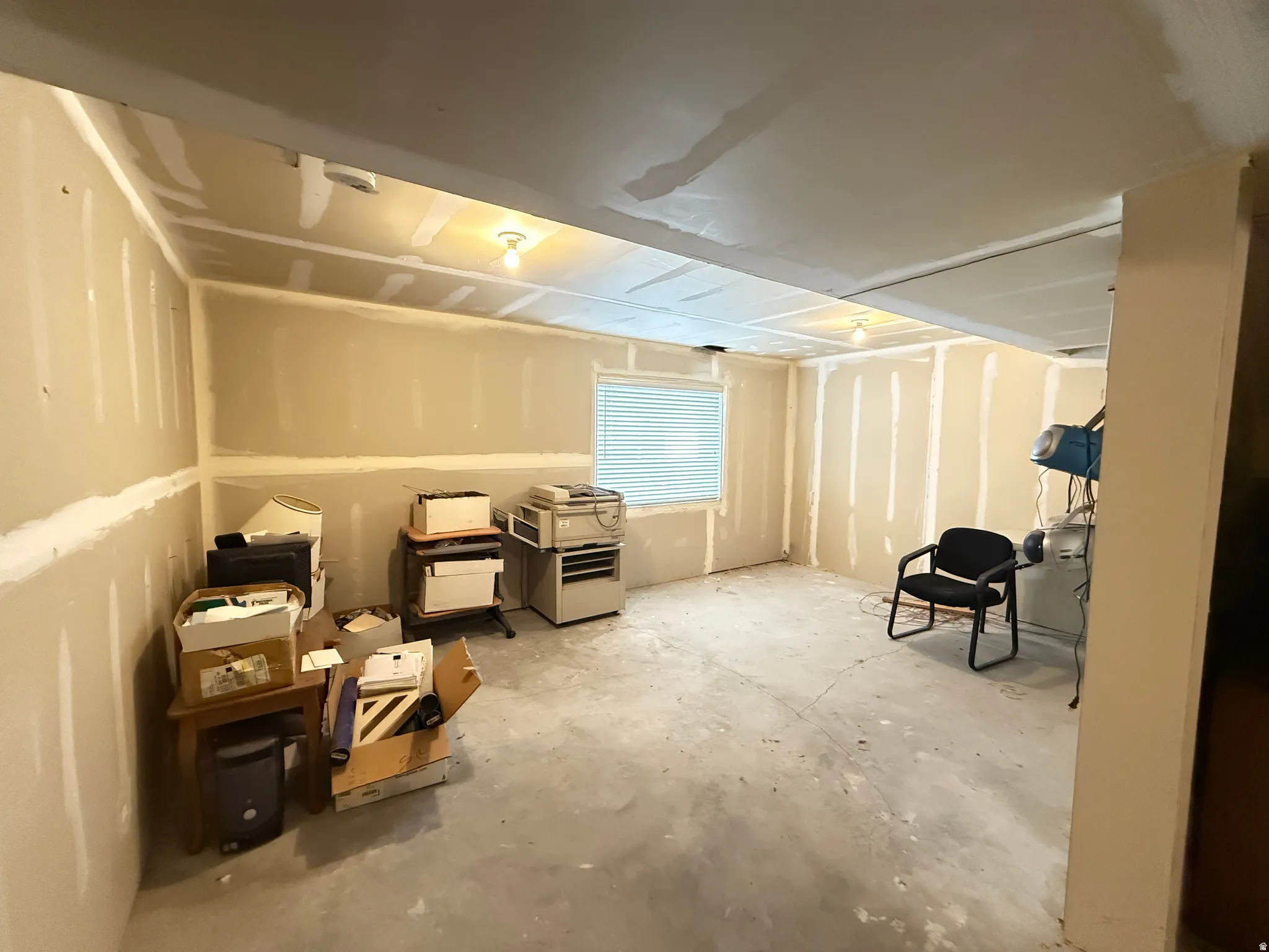 Unfinished interior space with exposed drywall, a single window with blinds, and overhead lighting
