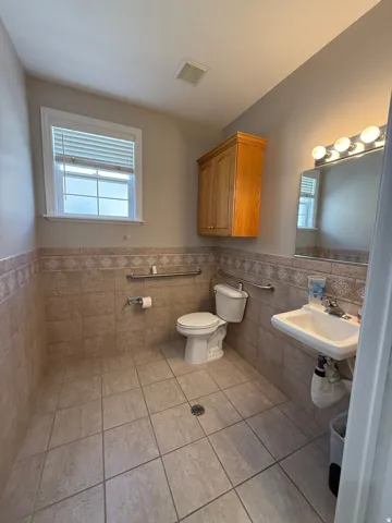 Tiled flooring with corresponding wainscoting, wall-mounted vanity sink, white window with blinds, and built-in wood-finish cabinetry