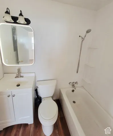 White vanity with an integrated sink and chrome faucet, white toilet, white bathtub with a wall-mounted showerhead, and wood-finish flooring
