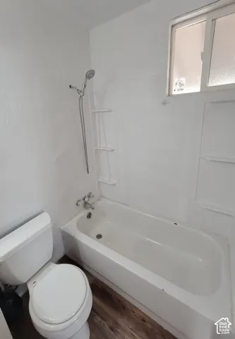 White bathtub with shower, white toilet, window with privacy glass, and wood-finish flooring