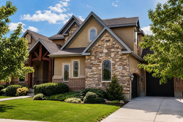 Stucco and stone facade with brick detailing, arched and rectangular windows, a prominent gable roofline, and an attached garage with dark doors