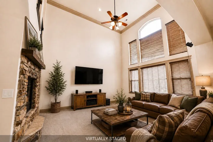 Vaulted ceiling with a ceiling fan, stone fireplace with a wood mantel, and tall windows with wood blinds
