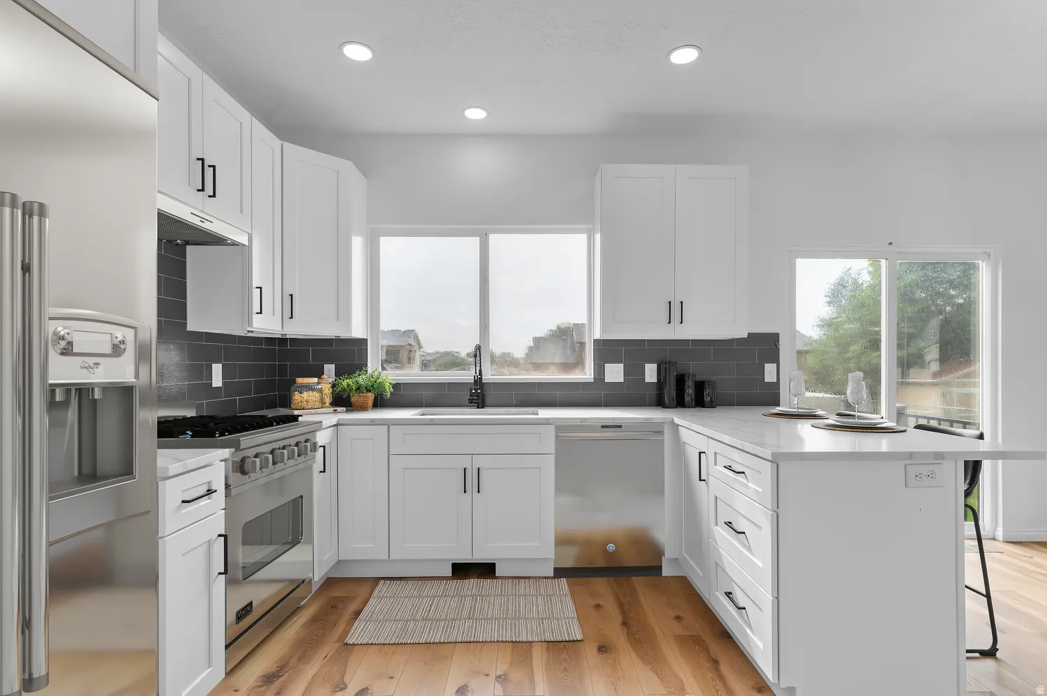Modern kitchen featuring white cabinetry with black hardware, stainless steel appliances, dark subway tile backsplash, light-toned countertops, and wood-finish flooring
