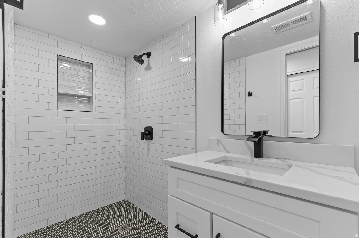 White subway tile shower surround with a built-in niche, black circular mosaic shower pan, white vanity with integrated sink, black wall-mounted faucet, and a rectangular black-framed mirror