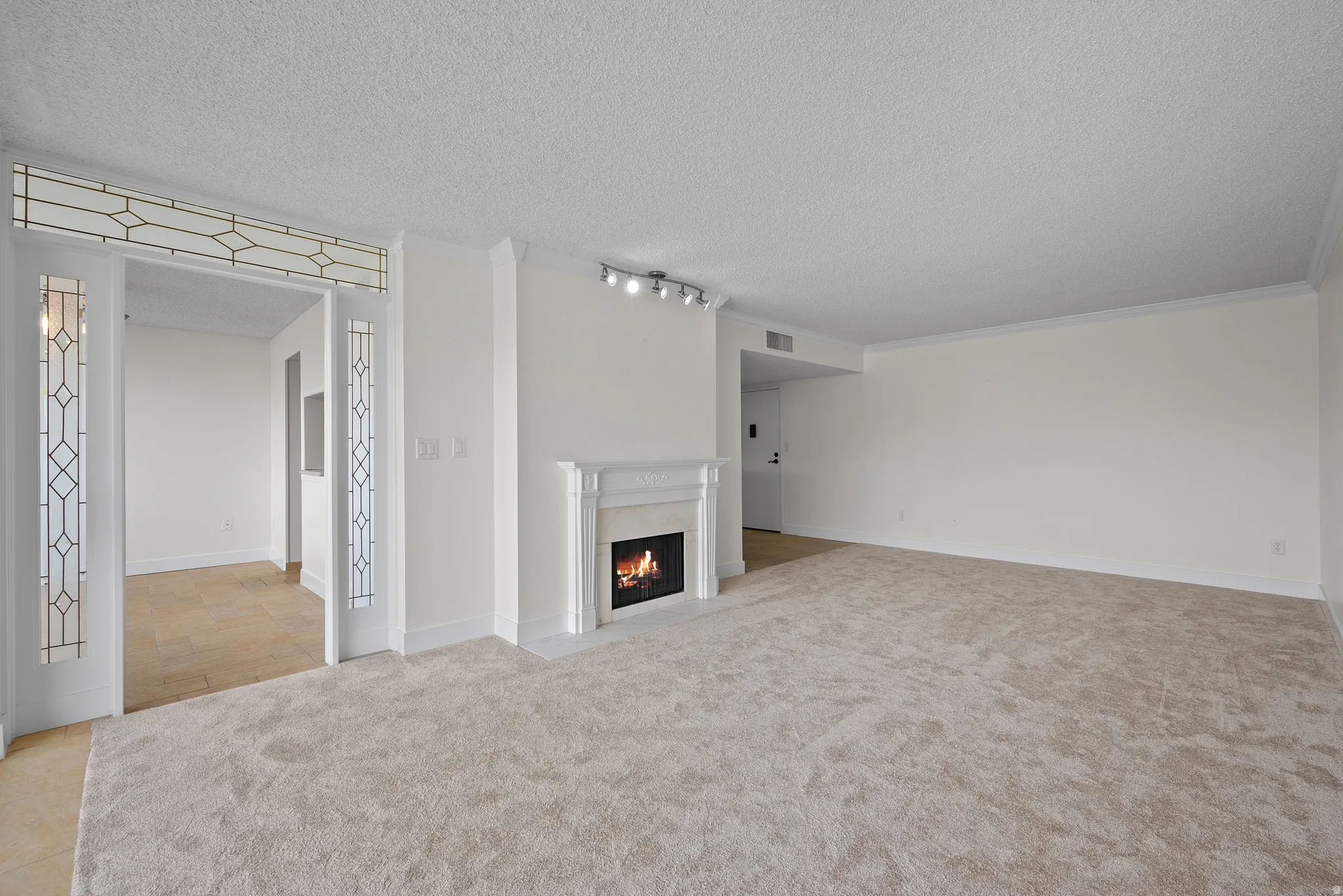 Spacious living area featuring light-colored carpeting, a fireplace with a traditional mantel, crown molding, and decorative glass panels framing an entryway