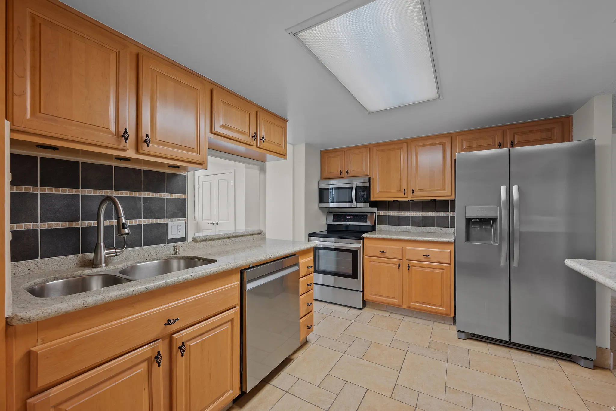 Neutral-toned tile flooring, light wood cabinetry, stainless steel appliances, dual basin sink, and mosaic tile backsplash