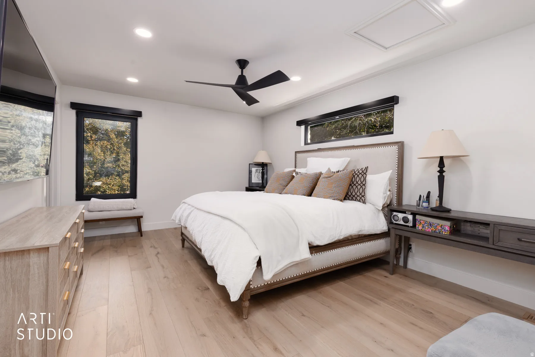 Light wood-finish flooring, recessed ceiling lights, an upholstered bed frame, a ceiling fan, and a transom window