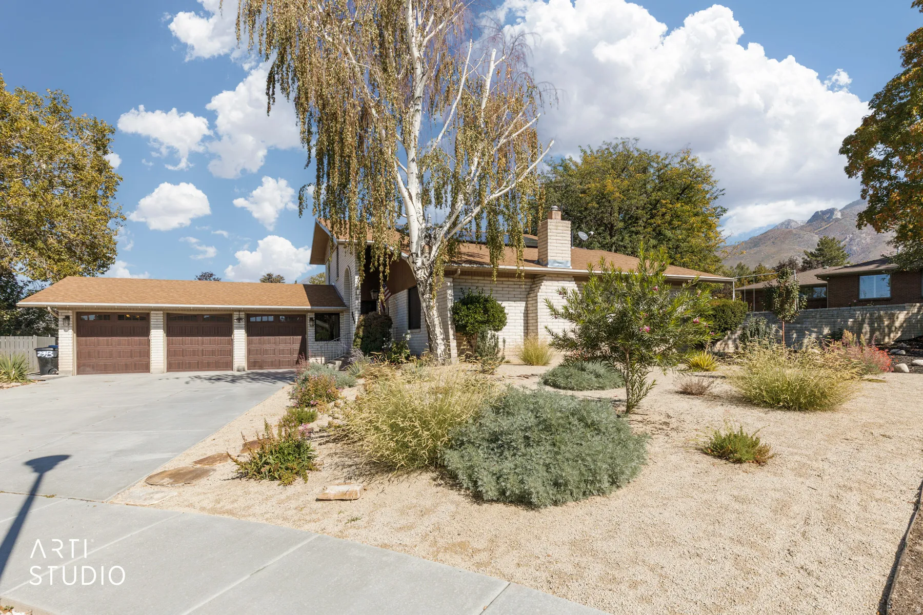 Three-car garage with individual doors, concrete driveway, xeriscaped front yard with varied plantings, and a prominent chimney feature