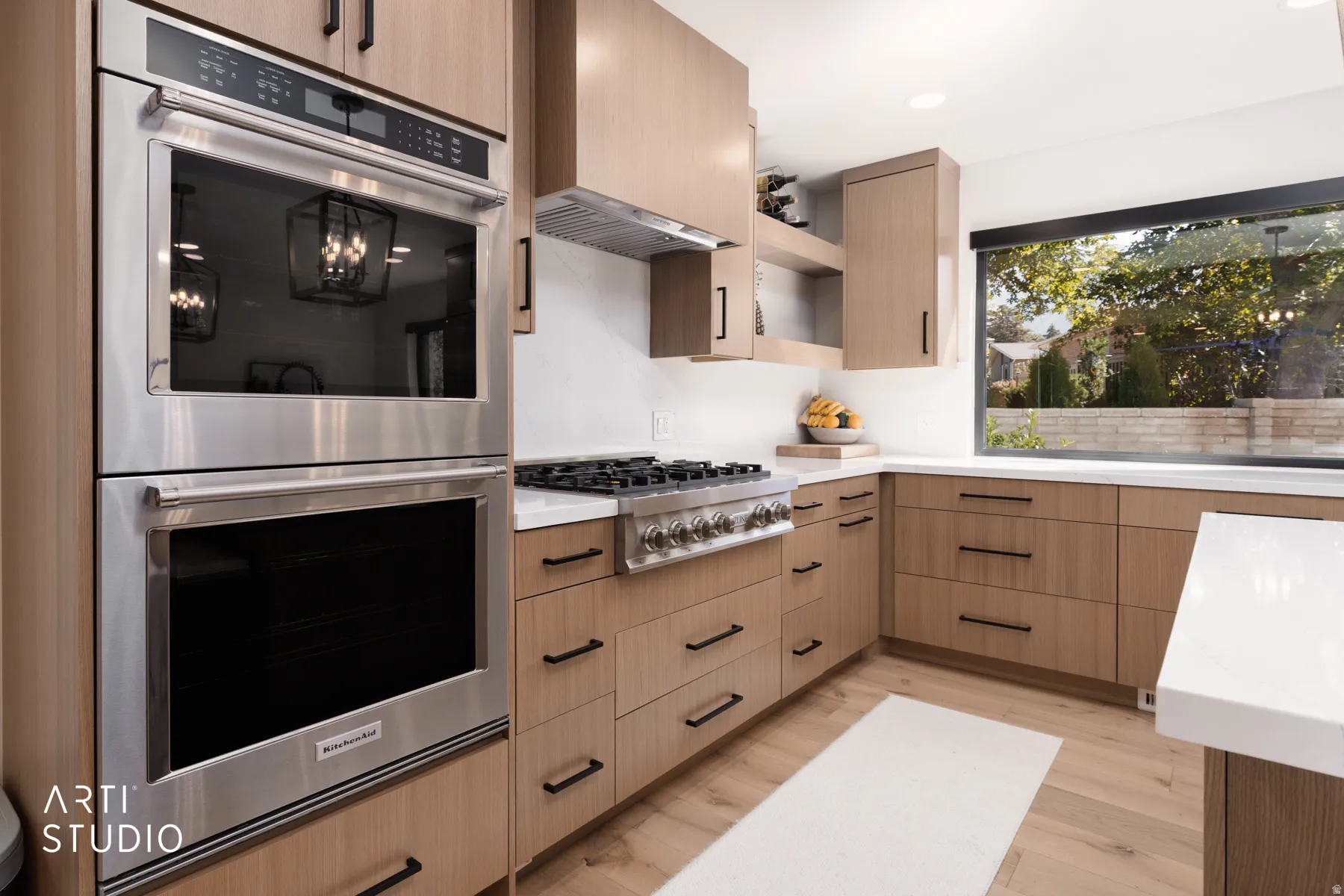 Stainless steel double oven, gas range, wood-finish cabinetry with black hardware, white countertops, and a large window