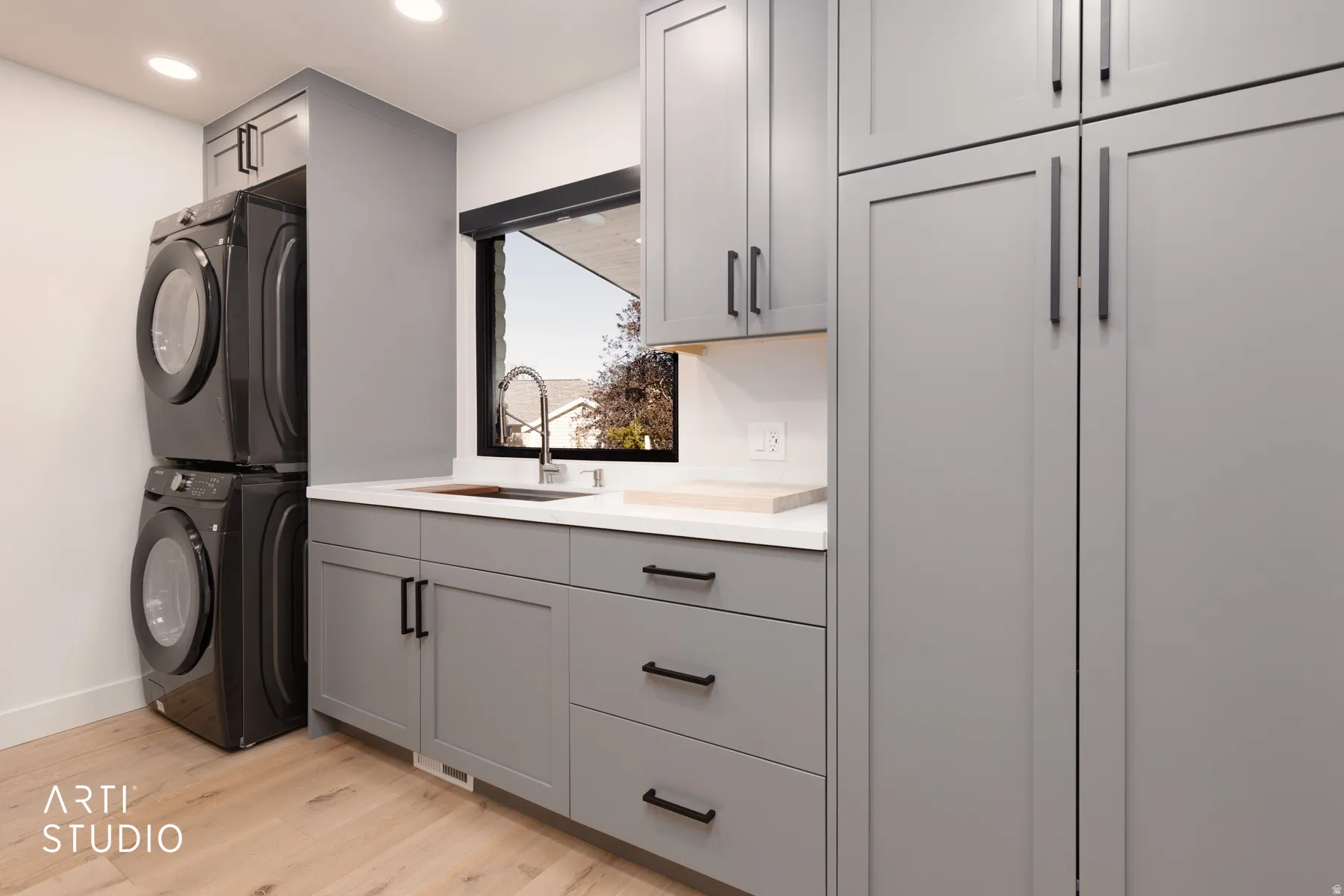 Integrated washer and dryer, light wood-finish flooring, white countertop, undermount sink with gooseneck faucet, and gray cabinetry with black hardware